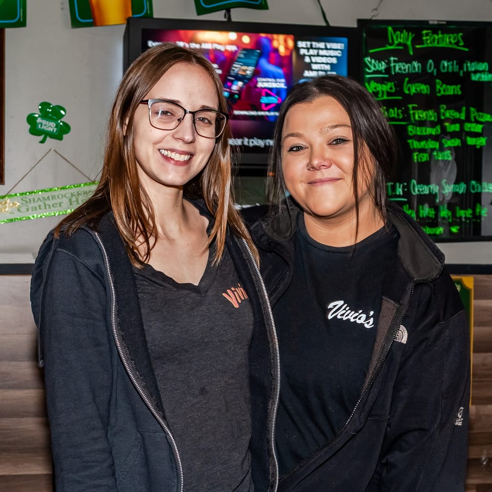 Two smiling Vivio's II staff members stand in front of a menu board and TV screen.