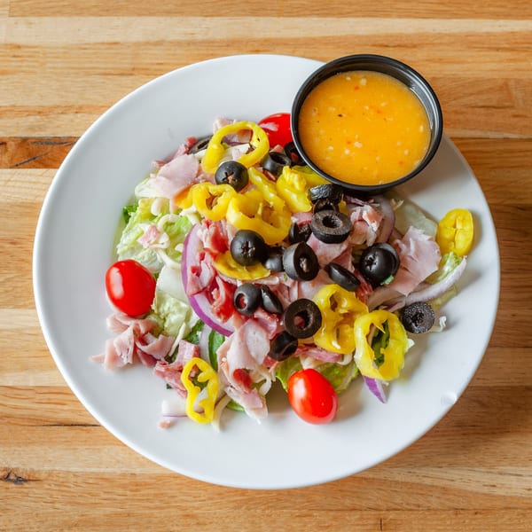 A large salad with ham, salami, olives, banana peppers, tomatoes, and red onion, with dressing.