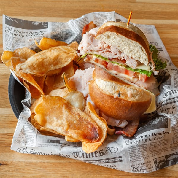 A Vivio's II turkey sandwich with cheese, lettuce, and tomato, served with potato chips on newspaper.