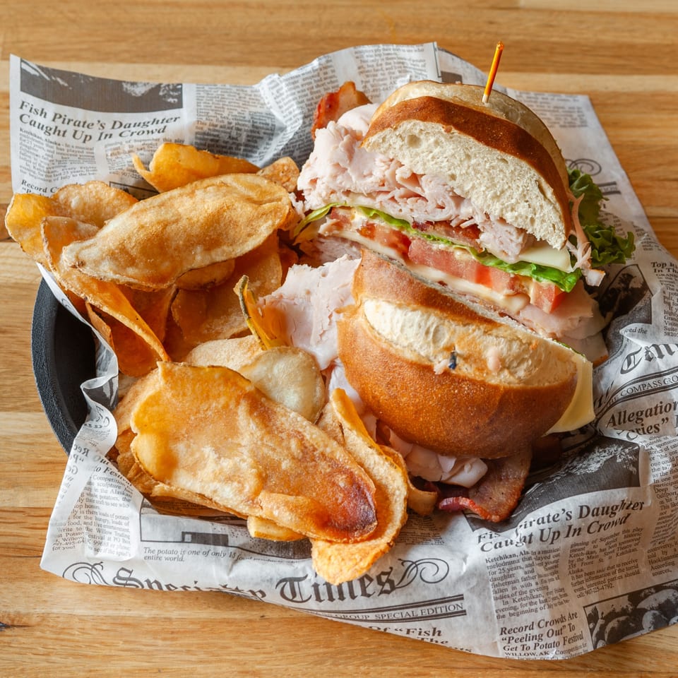 A Vivio's II turkey sandwich with cheese, lettuce, and tomato, served with potato chips on newspaper.